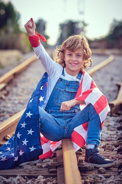 Corpo cheio de menino positivo envolto em bandeira colorida dos EUA olhando para a câmera com o braço levantado enquanto estava sentado em uma ferrovia de metal