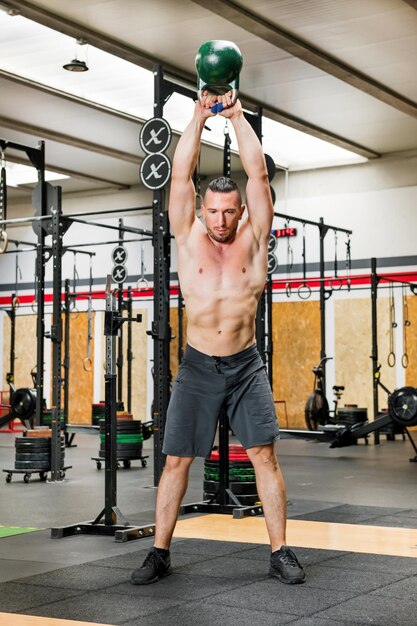 Corpo cheio de músculo atleta sem camisa em calções levantando pesado kettlebell sobre a cabeça enquanto trabalha no ginásio