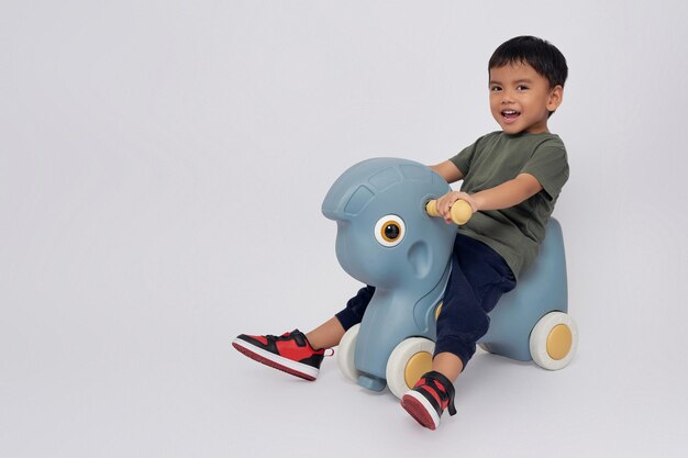 Foto corpo cheio pequeno sorridente feliz rapaz asiático de 23 anos vestindo camiseta verde sentado em um cavalo de balanço de brinquedo brincando com um brinquedo e olhando para a câmera isolado em fundo branco