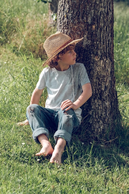 Corpo inteiro de criança descalça com cabelos cacheados e chapéu de palha, sentado na grama verde e apoiado no tronco de uma árvore enquanto relaxa em um dia de verão e desvia o olhar