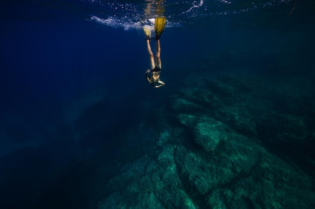 Corpo inteiro de jovem mergulhador sem camisa irreconhecível em nadadeiras amarelas nadando sob o mar azul profundo em direção ao recife de coral durante as férias de verão