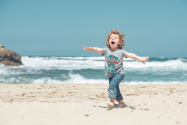 Corpo inteiro de menino ativo e brincalhão em roupas casuais correndo na costa arenosa perto do mar agitado enquanto se diverte na natureza