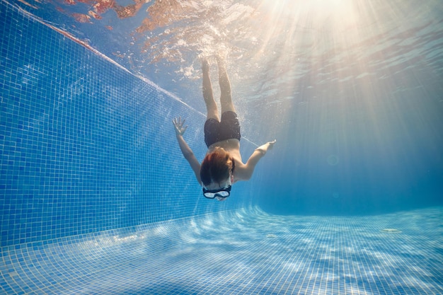 Corpo inteiro de menino descalço em óculos e shorts nadando na piscina limpa sob o sol