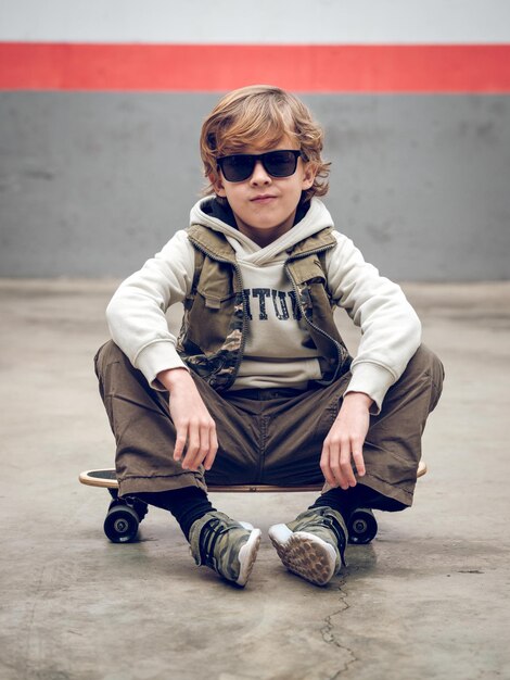 Foto corpo inteiro de menino sério com roupas elegantes e óculos de sol, sentado no skate no chão de concreto enquanto olha para a câmera