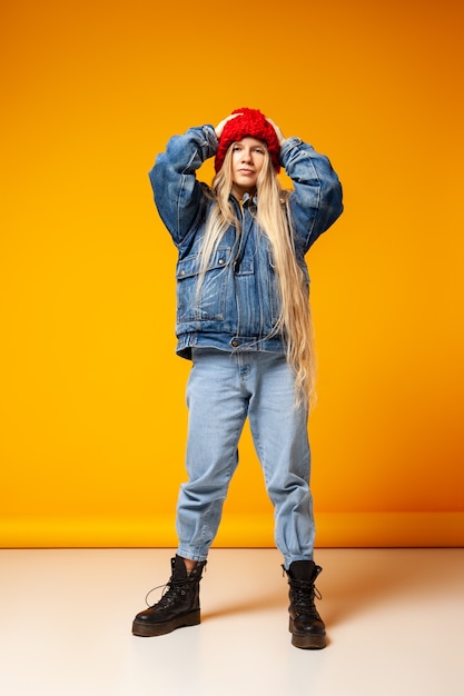 Corpo inteiro de modelo feminino milenar informal brincalhão em jaqueta jeans e jeans escondendo o rosto sob o chapéu de malha em pé contra um fundo laranja em estúdio