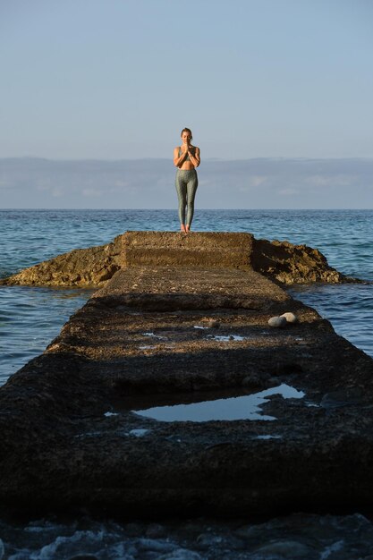 Corpo inteiro de uma jovem em forma em roupas esportivas praticando ioga em pose de montanha na costa rochosa e fazendo gesto namastê