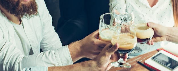 Corporativo. Manos de amigos, colegas durante beber cerveza, divertirse, tintinear botellas, vasos juntos.