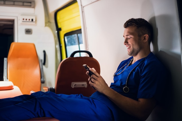 Corpsman em um uniforme azul, sentado em um carro de ambulância usando smartphone