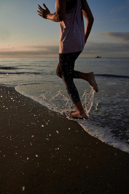 Foto corredor anônimo descalço em roupas esportivas correndo rápido na costa arenosa molhada ao longo do mar ao pôr do sol durante o treinamento na natureza