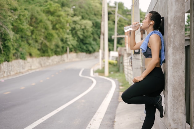 Corredor asiático novo da mulher que descansa após a corrida do exercício e a água potável na estrada da rua.