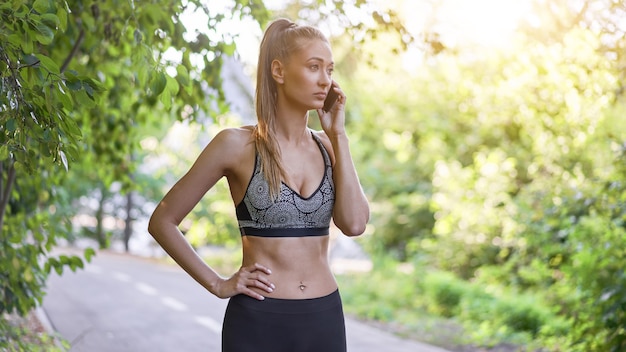 Corredor de mulher em pé antes do exercício parque de verão falando ao telefone.