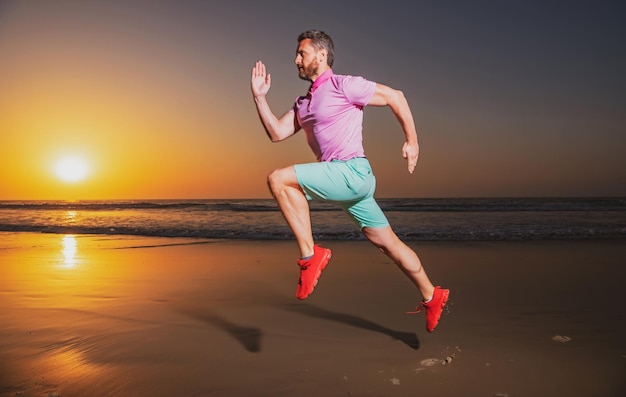 Corredor esportivo correndo na praia do mar de verão atraente homem em forma correndo no treino de luz do sol ao ar livre