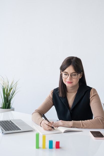 Corredor femenino elegante joven que mira el gráfico del cubo multicolor en el escritorio mientras analiza las finanzas de la empresa por lugar de trabajo