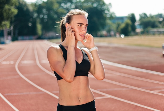 Corredor jovem alegre no sportswear está ouvindo música com fones de ouvido no estádio revestido de vermelho ao ar livre