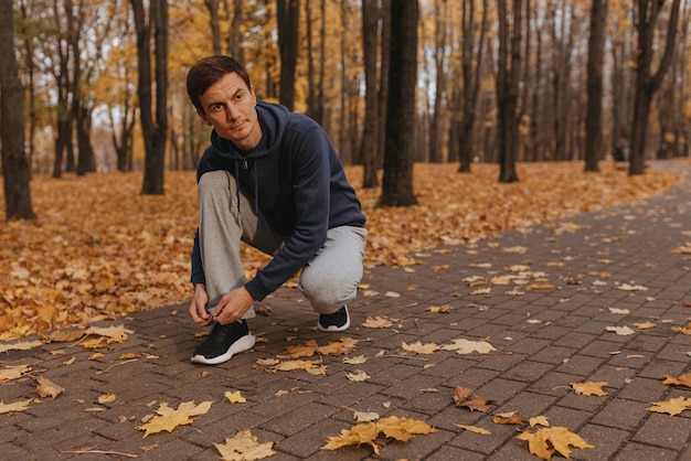 Corredor masculino atlético en ropa deportiva atando cordones de zapatos en zapatillas de deporte en la pasarela en el parque de otoño