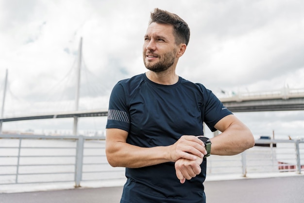 Corredor masculino enfocado chequeando el reloj de fitness contra el telón de fondo del puente urbano que encarna el estilo de vida activo