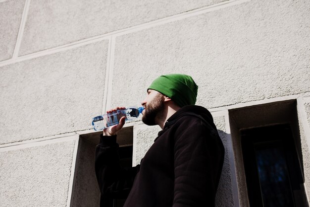 Corredor masculino sedento em roupas esportivas pretas e segurando uma garrafa de água se refrescar depois de correr, homem estiloso bebendo depois de um treino de treino na cidade