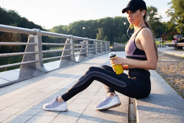 Corredor de la mujer joven descansando después de la sesión de entrenamiento en la mañana soleada. Modelo de fitness femenino sentado en la calle junto al estanque en la ciudad. Emparejador femenino tomando un descanso de la ejecución de ejercicios