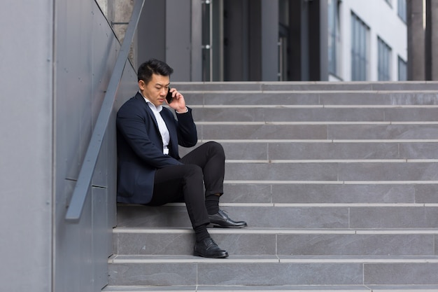 El corredor de negocios asiático informa malas noticias hablando por teléfono, sentado cerca de la oficina en las escaleras, deprimido, perdiendo la esperanza y con el trabajo en quiebra