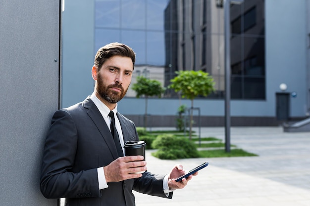 Corredor de negocios exitoso en un traje de negocios hablando por teléfono cerca de la oficina afuera, banquero sosteniendo una taza de café durante la pausa del almuerzo sonriendo