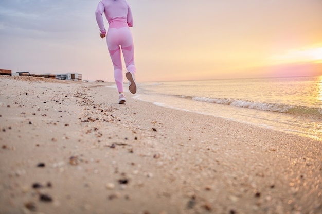 Foto corredora feminina com roupa esportiva rosa, praticando corrida na praia de manhã cedo