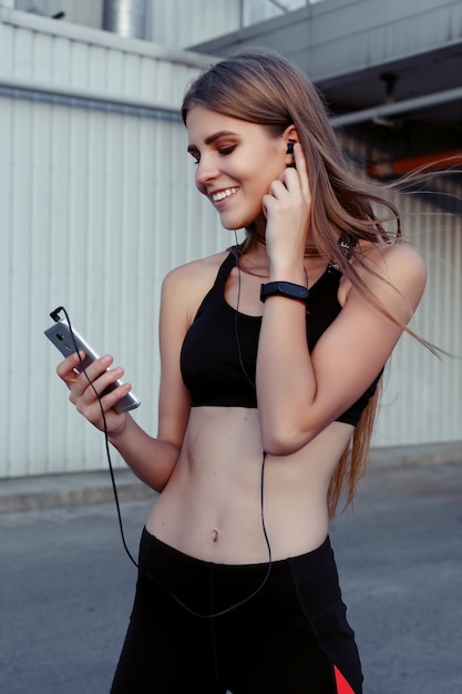 Corredoras tomando descanso después del entrenamiento en la ciudad. Mujer fitness en ropa deportiva con auriculares escuchando música y mirando a otro lado riendo.