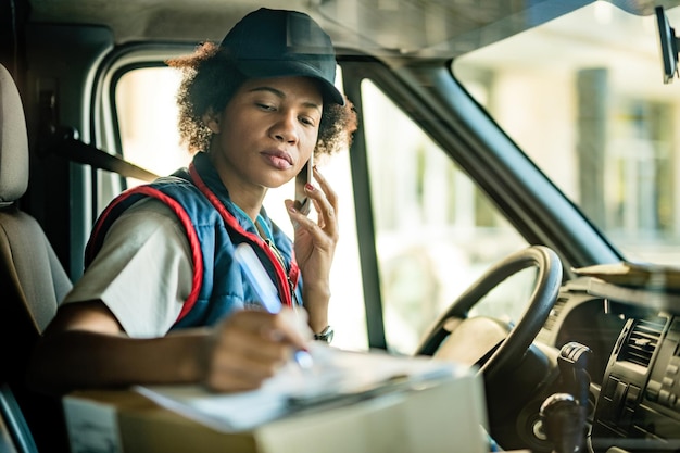 Correio afro-americano feminino tomando notas enquanto fala ao telefone em uma van de entrega