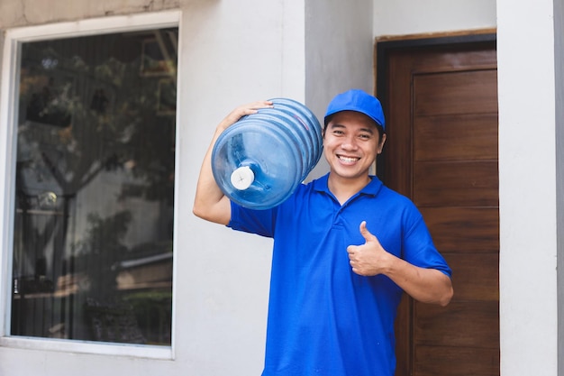 Correio asiático alegre carrega galão de água no ombro, traz o pedido ao cliente e mostra o polegar para cima