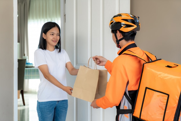 Correio asiático do homem na bicicleta que entrega o alimento no sorriso uniforme alaranjado e que prende o saco do alimento na casa dianteira e a mulher asiática que aceita uma entrega das caixas do entregador.