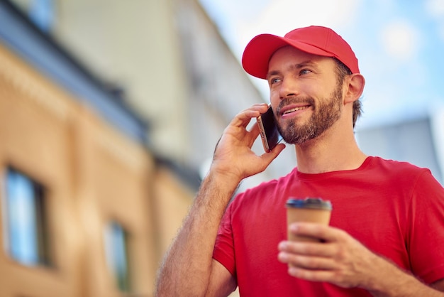 Foto correio caucasiano jovem segurando a xícara de café e falando no telefone
