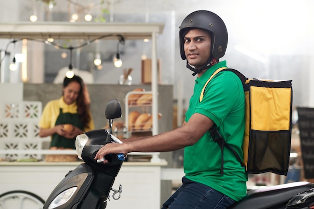 Correio com grande sacola isolada nas costas sentado em uma scooter pronto para entregar o pão da padaria local