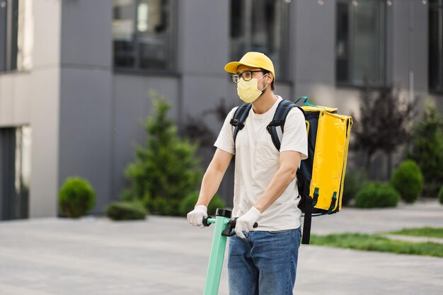 Correio de entrega de comida usando mochila térmica de máscara facial amarela anda de scooter elétrico