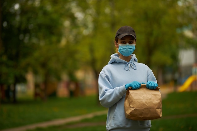 Correio de menina em uma máscara médica e luvas médicas na rua Jovem trabalhadora de entrega de alimentos