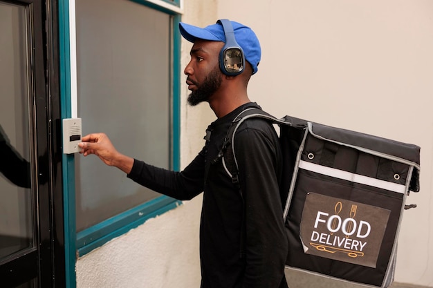 Correio de serviço de entrega tocando vista lateral da campainha do prédio, esperando o cliente ao ar livre. Entregador afro-americano com mochila térmica em frente à porta, entregando almoço para viagem