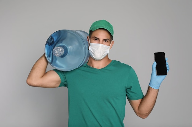 Correio em máscara médica com garrafa para bebedouro mostrando celular em fundo cinza claro Entrega durante a quarentena de coronavírus