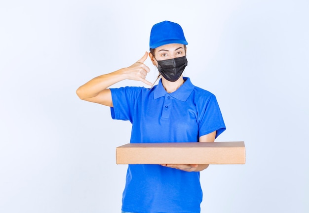 Correio feminino de uniforme azul e máscara facial, segurando uma caixa de papelão e pedindo uma ligação.