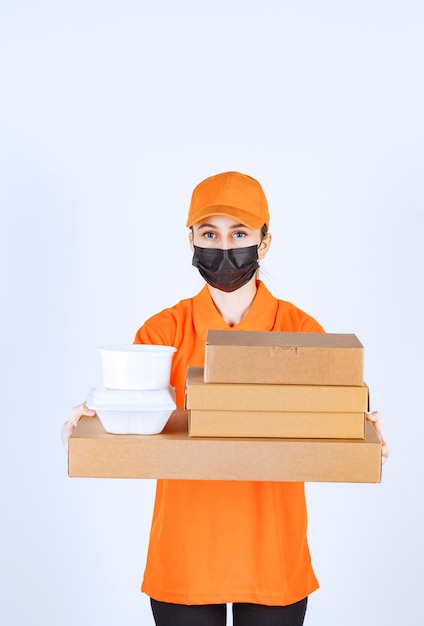 Correio feminino em uniforme amarelo e máscara preta segurando vários pacotes de papelão e caixas para viagem.