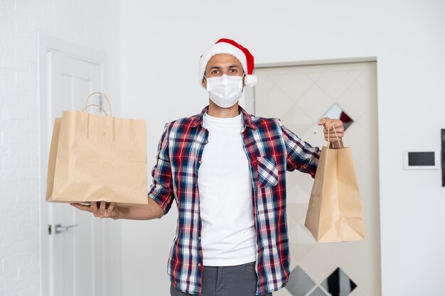 Correio masculino com máscara médica protetora e chapéu de Papai Noel segurando a caixa de papelão. pedido de mercadorias e entrega em quarentena.