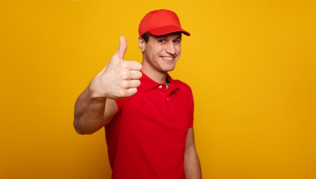 Correio ou entregador feliz jovem bonito com uniforme vermelho está posando isolado em fundo amarelo.