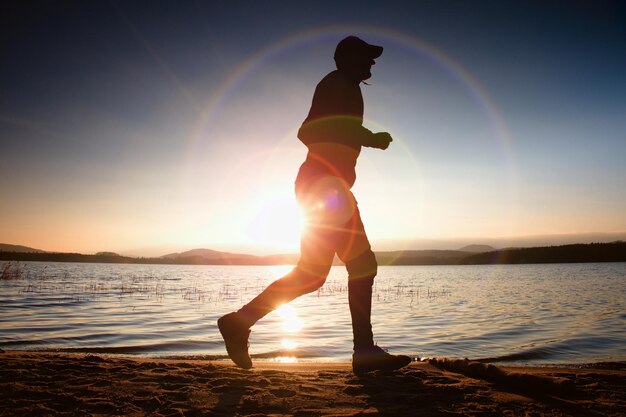 Foto correndo homem em raios de sol na praia desportista em boné de beisebol correndo durante o nascer do sol acima do mar