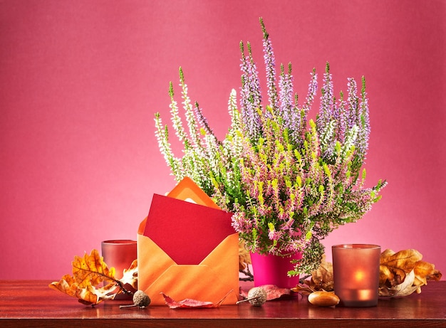 Correo de otoño. Naturaleza muerta con tarjeta de felicitación, sobre, flores de brezo en maceta, hojas secas y velas