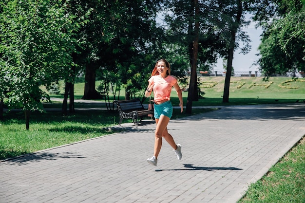 Correr a una joven deportista sonriente en el parque con ropa deportiva en el fondo de árboles verdes en baldosas grises en un día soleado de verano