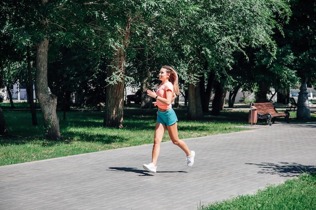 Correr a una joven deportista sonriente con reloj y cabello que fluye en el parque con camiseta rosa, pantalones cortos verdes y entrenadores blancos sobre árboles verdes de fondo y hierba en un día soleado y ventoso de verano desde el frente