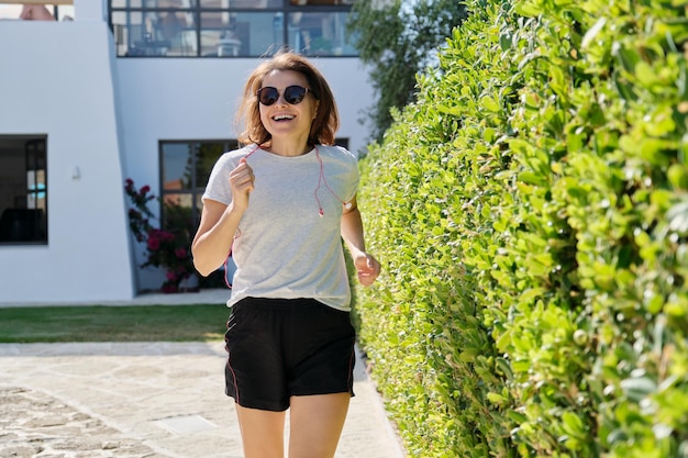 Correr sonriente mujer fitness de mediana edad en un día soleado de verano. Estilo de vida activo y saludable en mujeres maduras.