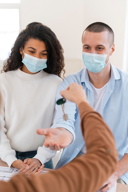 Corretora de imóveis entregando casal com máscaras médicas e as chaves de sua nova casa