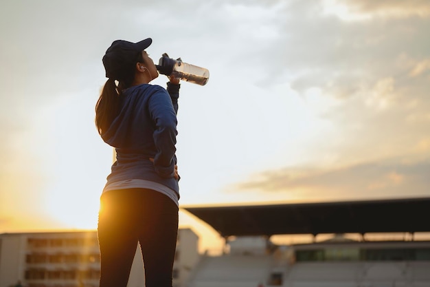 Corrida, fitness, corrida, exercício, estilo de vida, conceito saudável. Mulheres jovens aquecem o corpo. Sente-se e beba um descanso de proteína de soro de leite depois de correr na pista de corrida ao redor do campo de futebol.