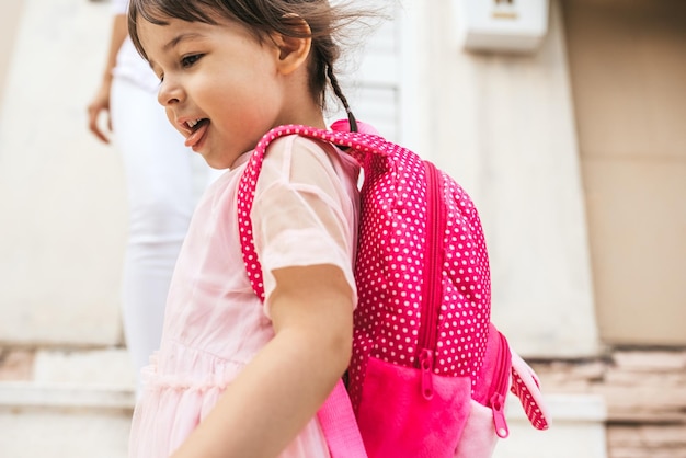 Cortada a imagem de close-up de linda criança mostrando a língua e sorrindo enquanto ela sai para o jardim de infância contra sua mãe Linda menina feliz usa mochila indo para o conceito de pessoas de educação pré-escolar