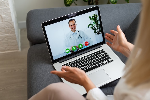 Cortada a vista traseira de uma jovem na sala de luz brilhante dentro de um apartamento aconchegante usando o laptop do pc. Ela conversando com um bom médico pela Internet