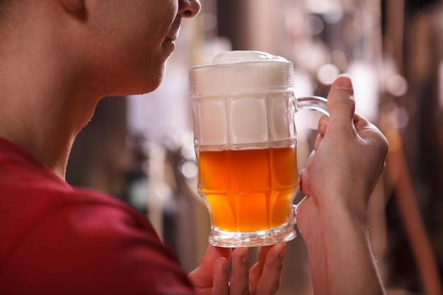 Cortado de perto de um cervejeiro profissional sorrindo, cheirando cerveja recém-fabricada em uma caneca