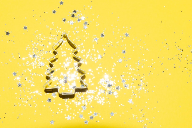 Foto cortador de galletas con temática navideña plateada sobre un fondo amarillo brillante con purpurina plateada y confeti de estrellas